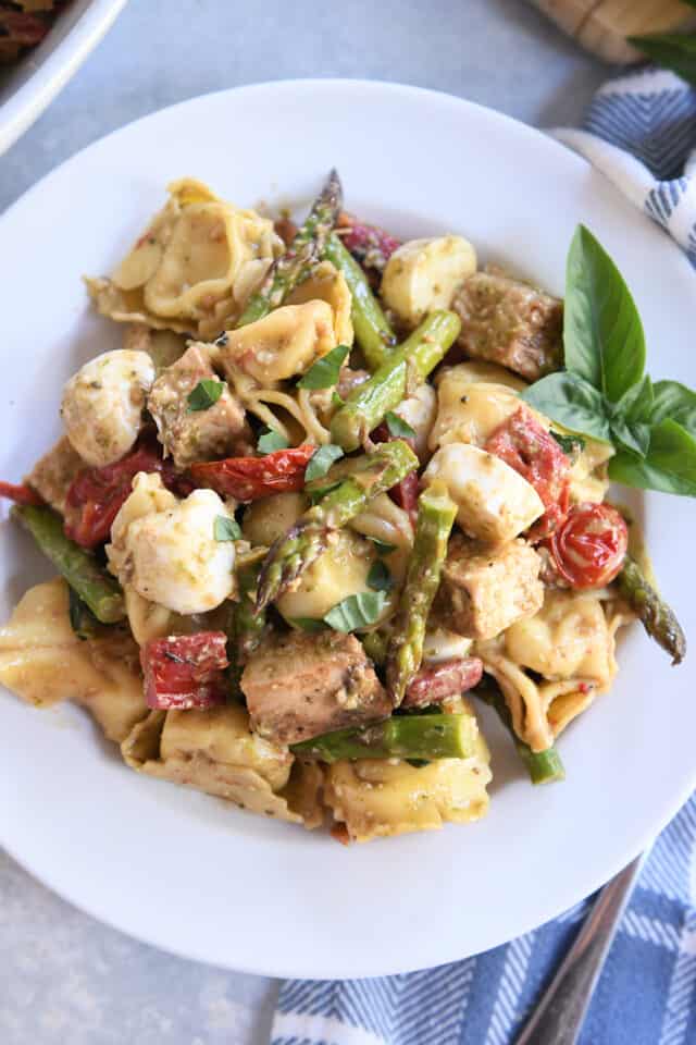 White plate with tortellini salad, fresh mozzarella, grilled chicken, and fresh basil.