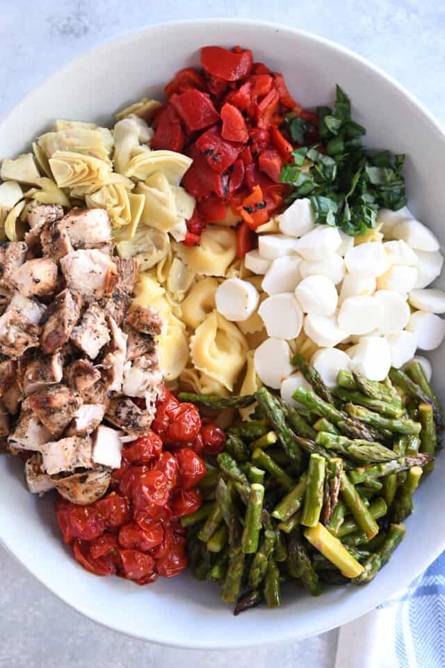 White bowl with artichokes, roasted red peppers, fresh basil, fresh mozzarella, asparagus, roasted tomatoes, grilled chicken and tortellini.