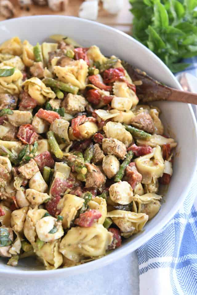 White bowl with wooden spoon on the side of tortellini salad with mozzarella, chicken, roasted red peppers and tomatoes.