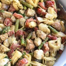 White bowl with wooden spoon on the side of tortellini salad with mozzarella, chicken, roasted red peppers and tomatoes.