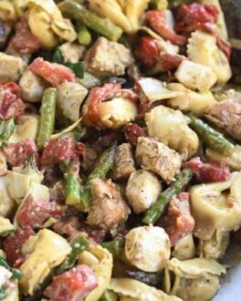 White bowl with wooden spoon on the side of tortellini salad with mozzarella, chicken, roasted red peppers and tomatoes.