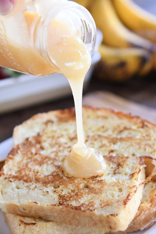 Pouring cream colored syrup over two pieces of French toast on white plate.