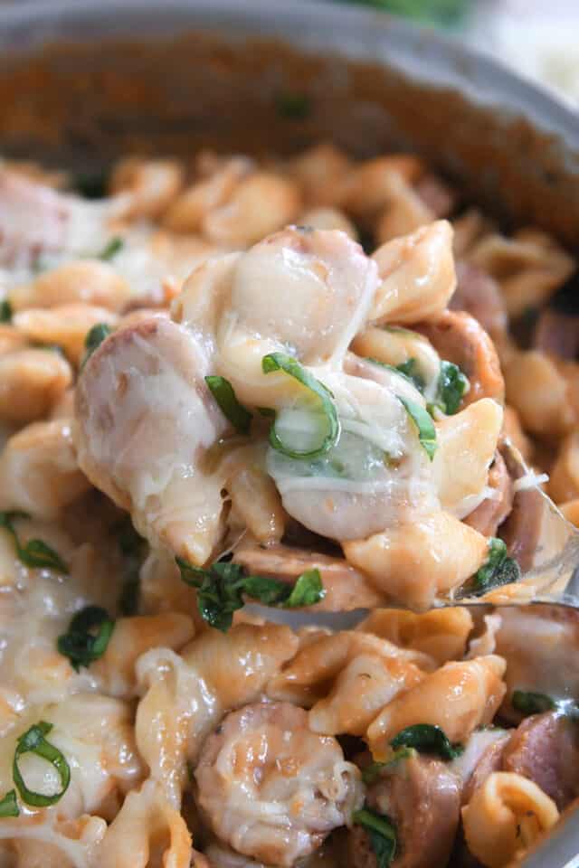 Spoon scooping up serving of pasta shells, fresh basil, sausage and cheese.