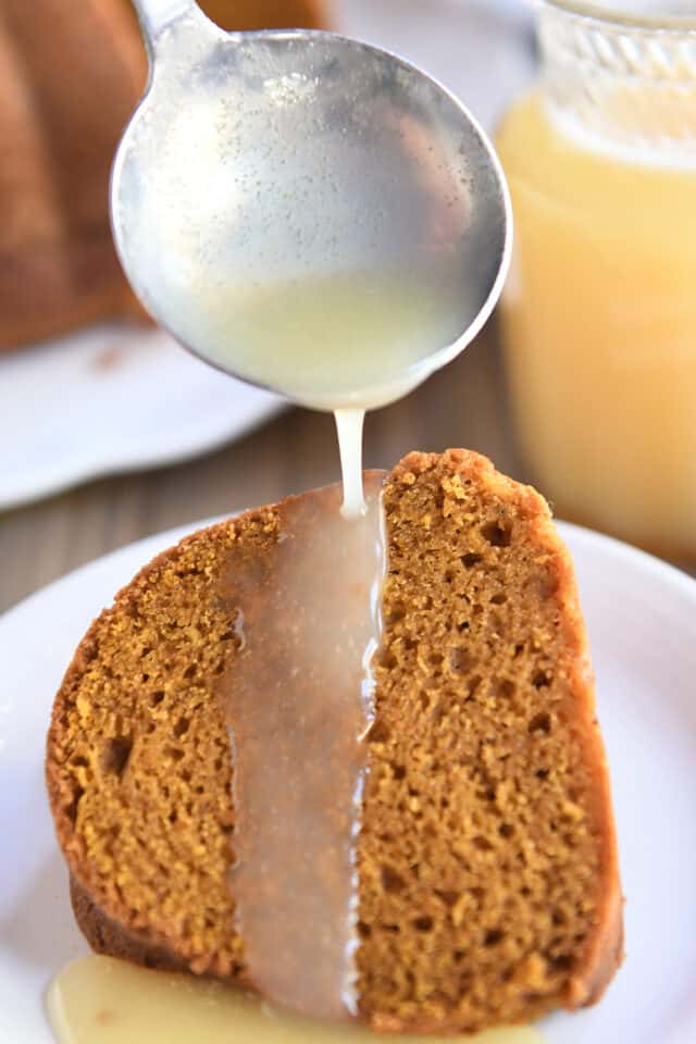 Small ladle spooning vanilla sauce over slice of pumpkin bundt cake on white plate.
