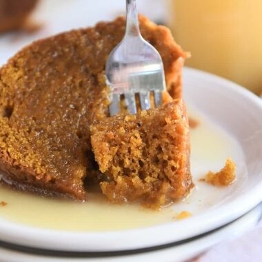 Bite of orange pumpkin cake being scooped out of big slice with fork on white plate.