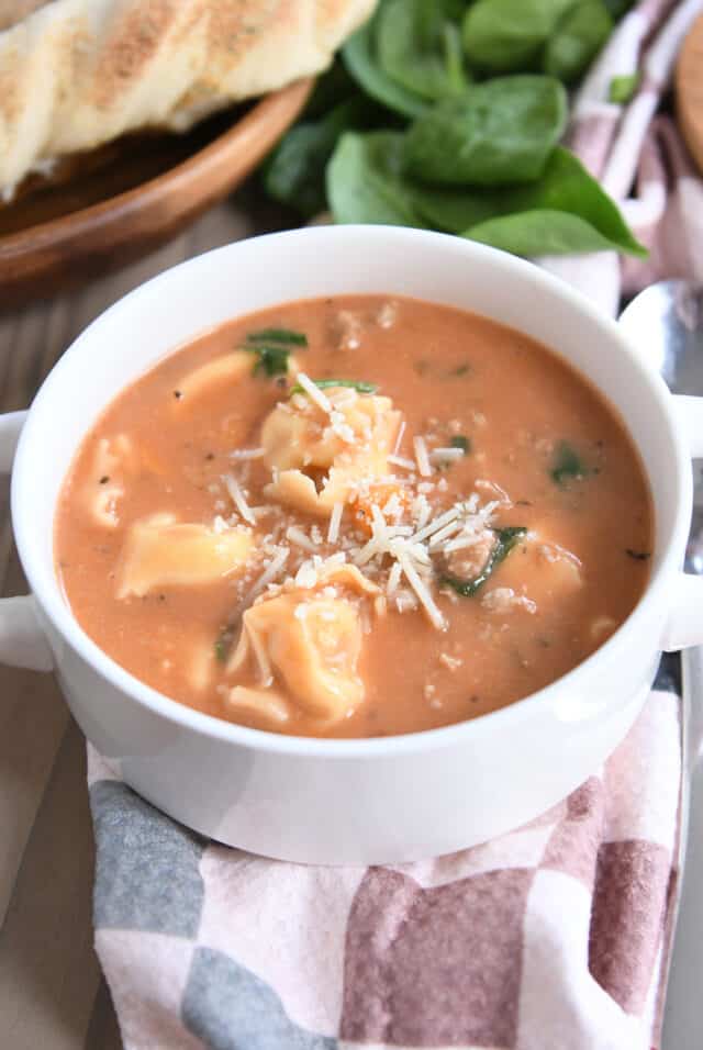 White bowl with handles filled with creamy tomato broth, tortellini, carrots, celery and Parmesan cheese.