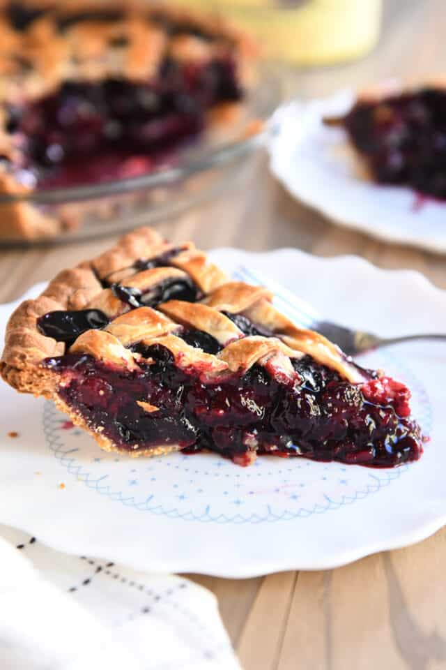 One slice of triple berry pie on a white scalloped plate with a fork.