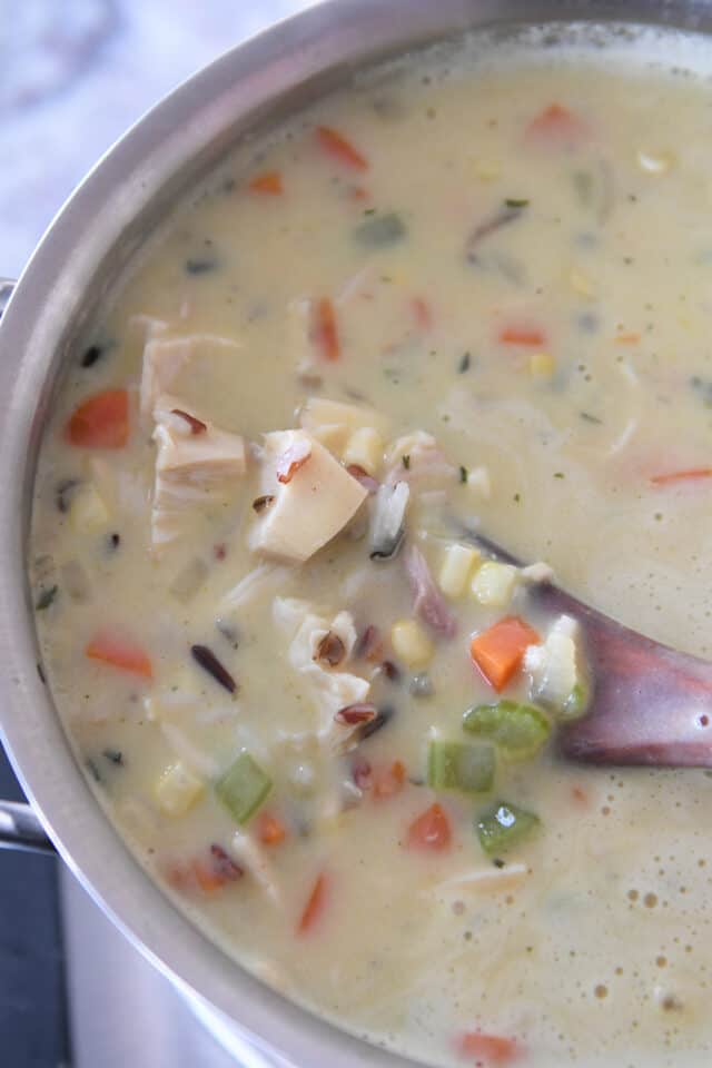Wooden spoon stirring soup with rice, chicken, carrots and celery in saucepan.