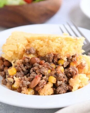 Portion of cornbread topped beef and bean casserole on white plate with fork.