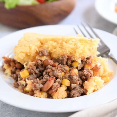 Portion of cornbread topped beef and bean casserole on white plate with fork.