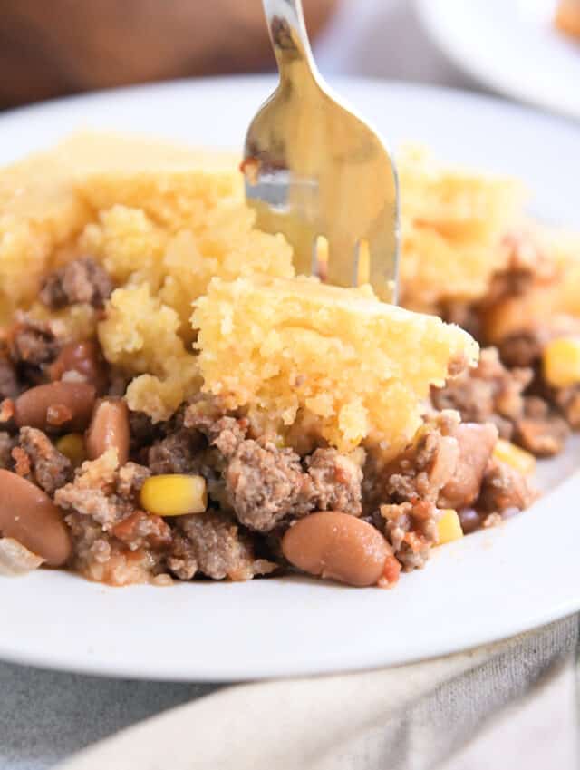 Fork taking bite of cornbread topped beef and bean casserole.