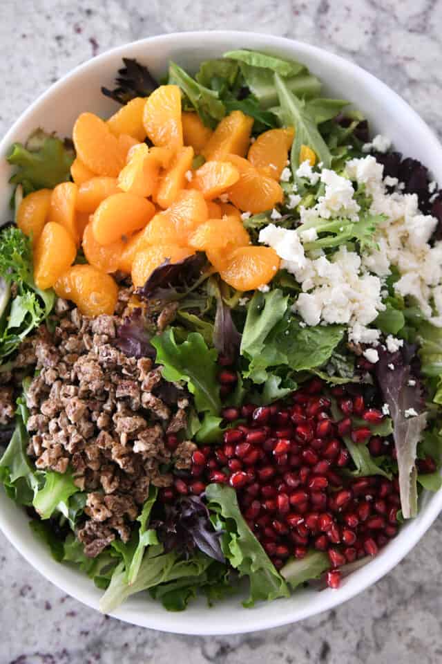 White salad bowl filled with salad greens, mandarin oranges, feta cheese crumbles, candied pecans and pomegranate arils.