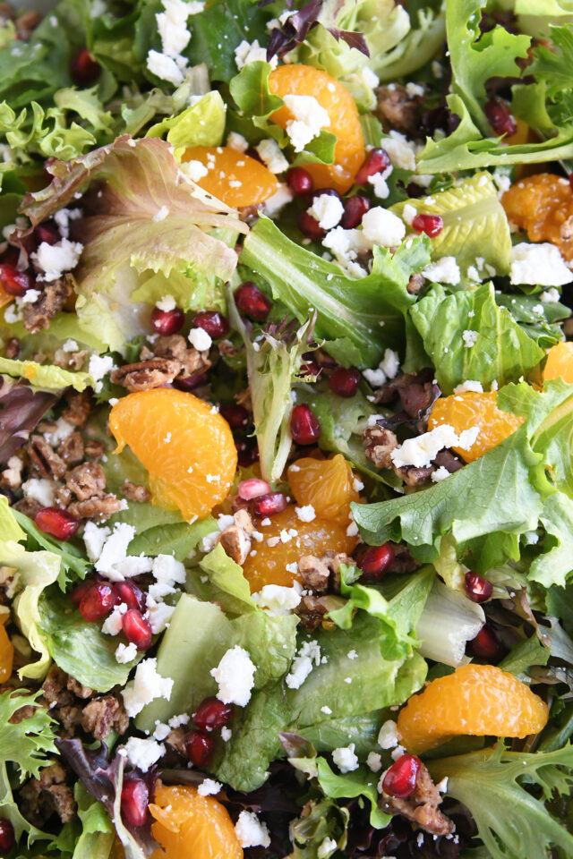 Close up view of salad greens, mandarin oranges, pomegranate arils, feta cheese and candied nuts.