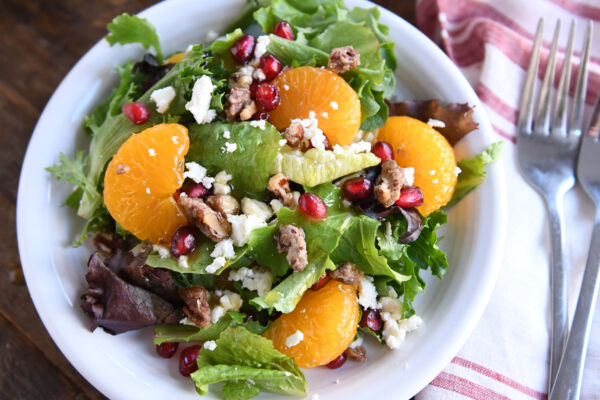 Salad greens, mandarin oranges, pomegranate arils, candied nuts and feta cheese on white plate.