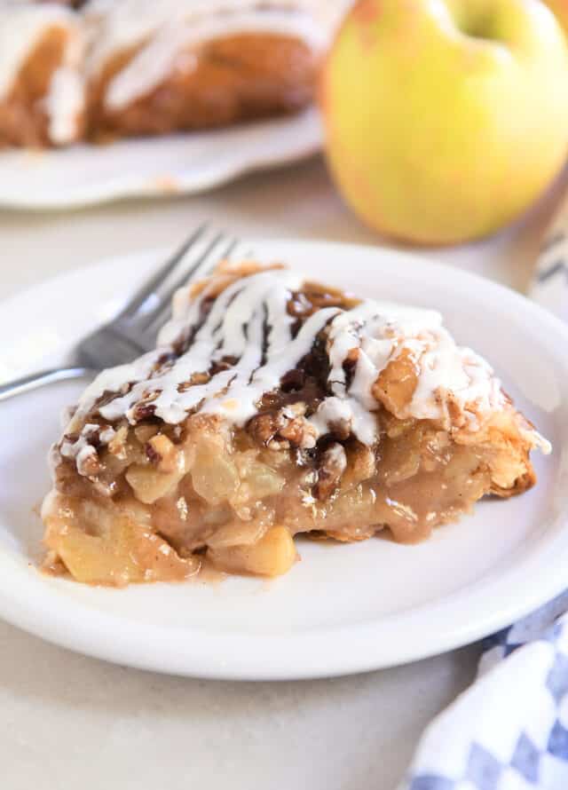Slice of apple pie on white plate with pecans and glaze on top.
