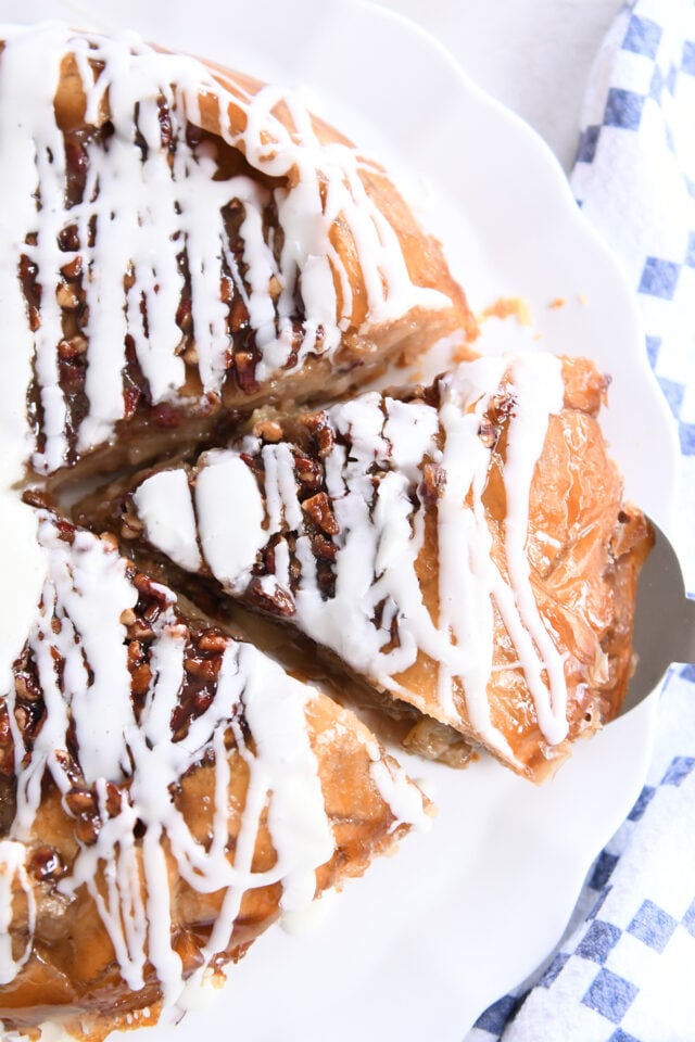 Slice of apple pie on white platter with pecans and glaze on top.