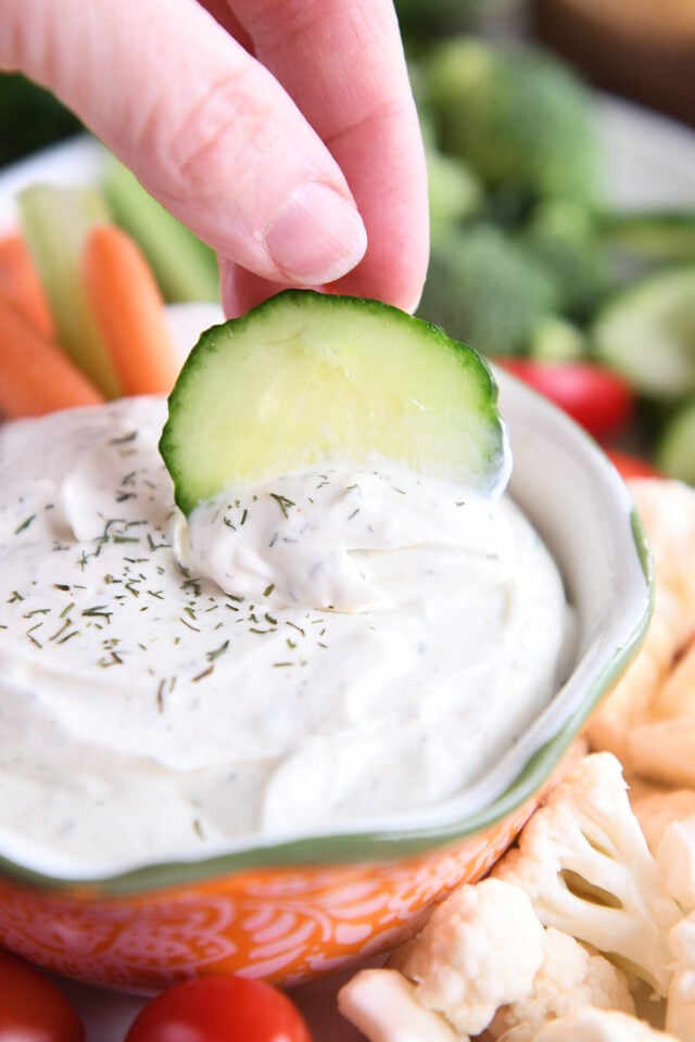 Dipping cucumber slice into veggie dip in orange bowl.