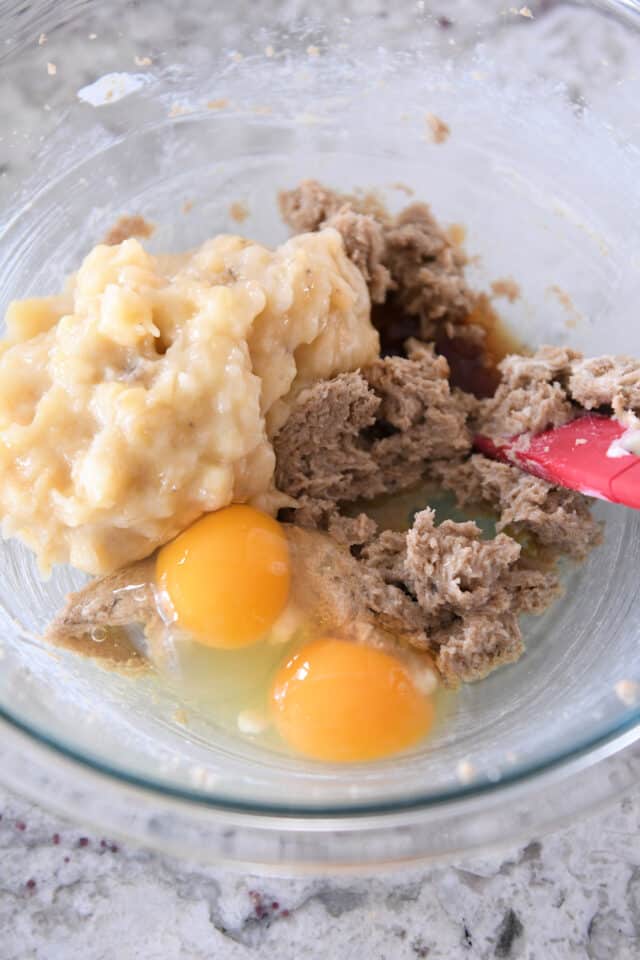 A glass bowl with mashed bananas, eggs, brown sugar and butter.