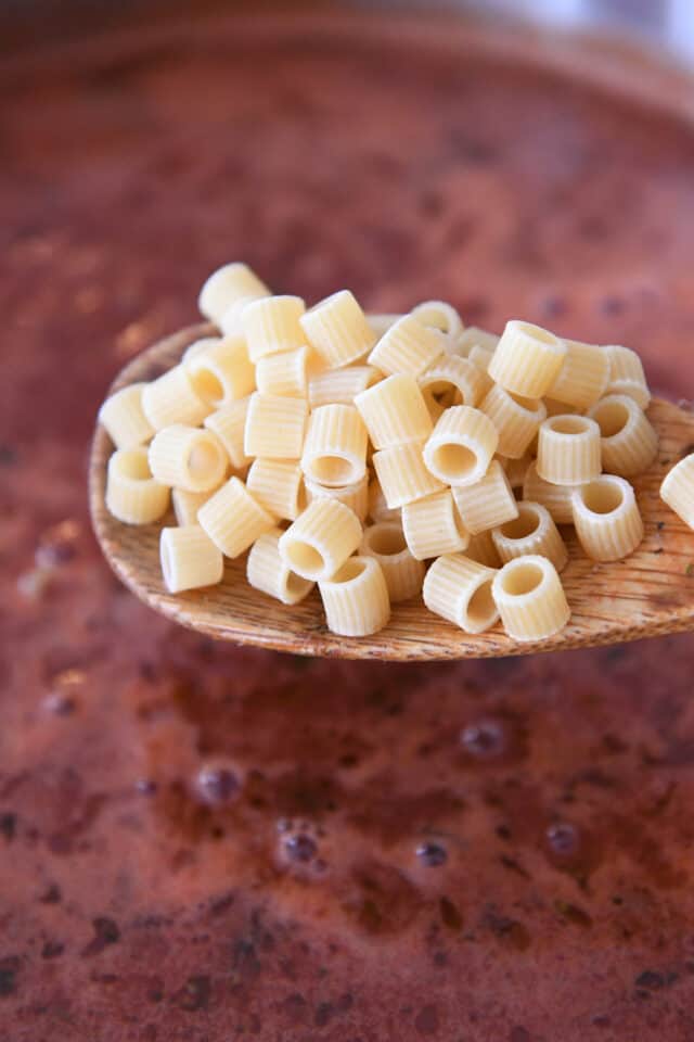 Ditalini noodles on wooden spoon above saucepan with tomato broth.