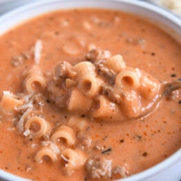 Spoon scooping up bite of soup with tomatoes, noodles, and ground beef.