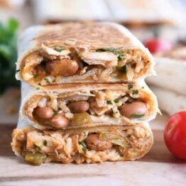 Three halves of crispy chicken wraps stacked on wooden board with cherry tomatoes.