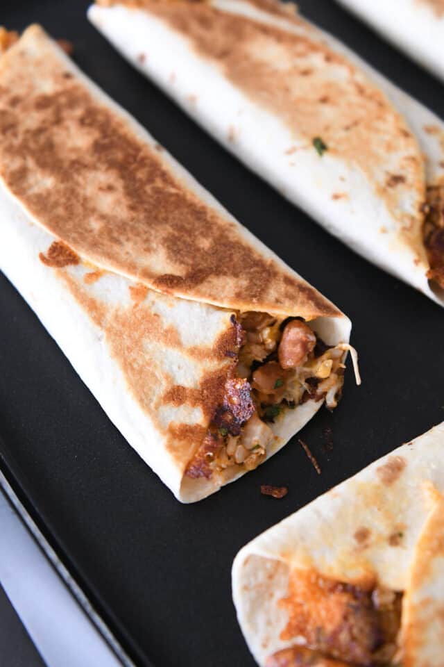 Tortilla wraps filled with beans, chicken, and rice are cooking on a nonstick skillet.