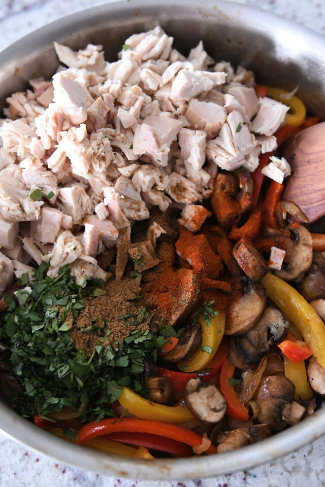 Skillet filled with sautéed peppers, mushrooms, coriander, cumin, chili powder and cooked chicken.