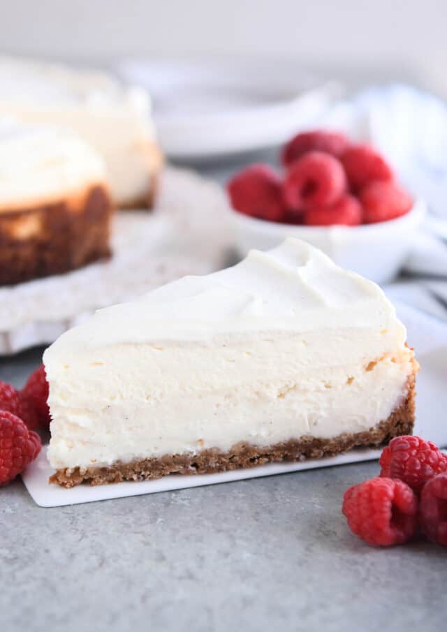 Slice of vanilla bean cheesecake on white serving spatula with raspberries.