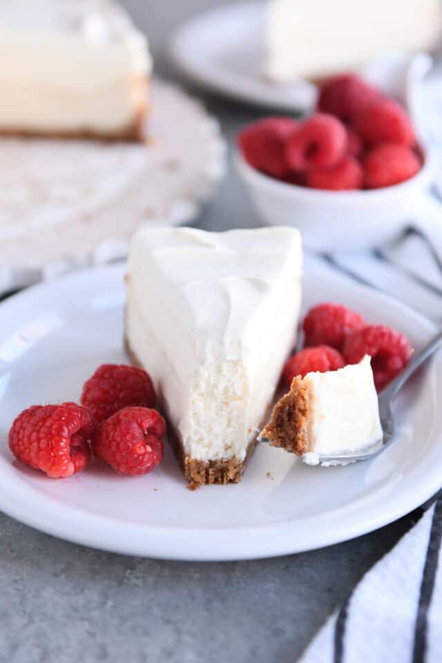 Bite of cheesecake on fork with white plate with raspberries.