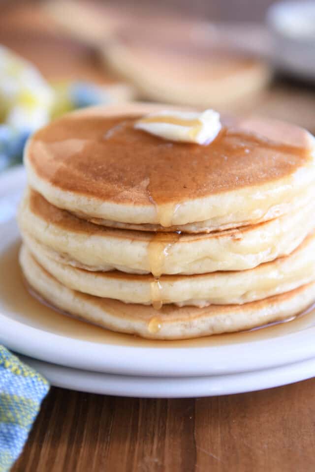 Stack of four pancakes on white plate with syrup dripping down side.