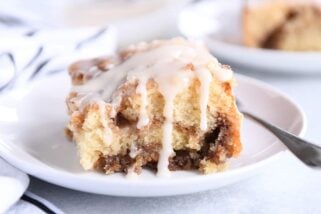 Square of cinnamon roll cake on white plate with glaze on top and sides.