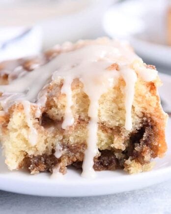 Square of cinnamon roll cake on white plate with glaze on top and sides.