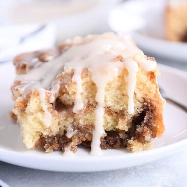 Square of cinnamon roll cake on white plate with glaze on top and sides.