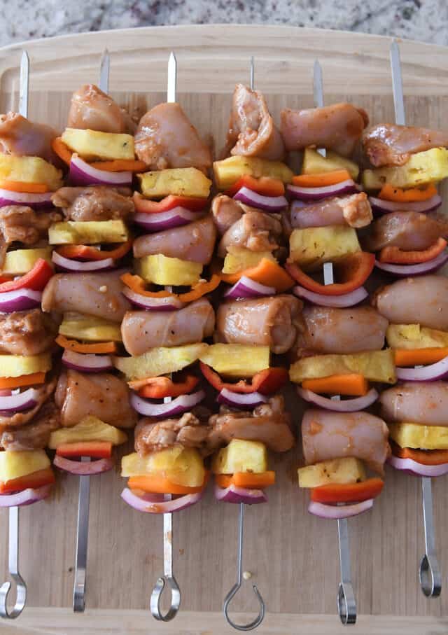 Six chicken, pineapple, veggie skewers on metal sticks on wood cutting board.