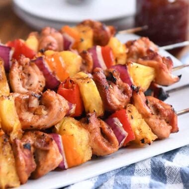 Several chicken, pineapple and pepper skewers on metal sticks on a white plate.