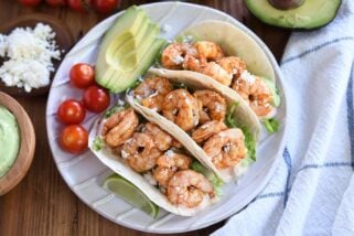 Three shrimp tacos on white ridged plate with tomatoes and avocado.