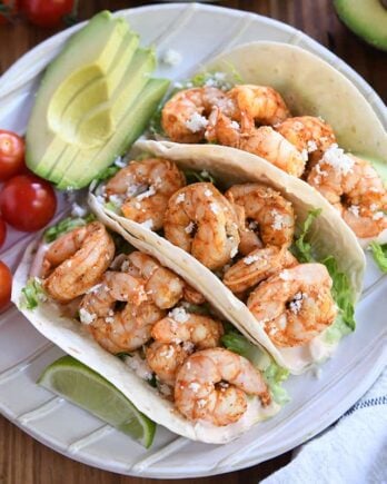 Three shrimp tacos on white ridged plate with tomatoes and avocado.
