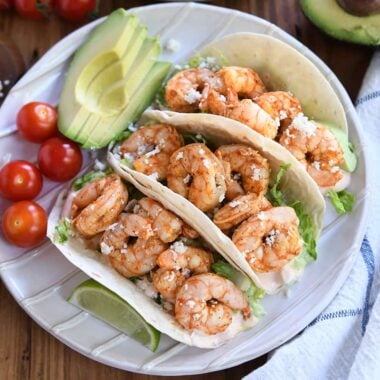 Three shrimp tacos on white ridged plate with tomatoes and avocado.