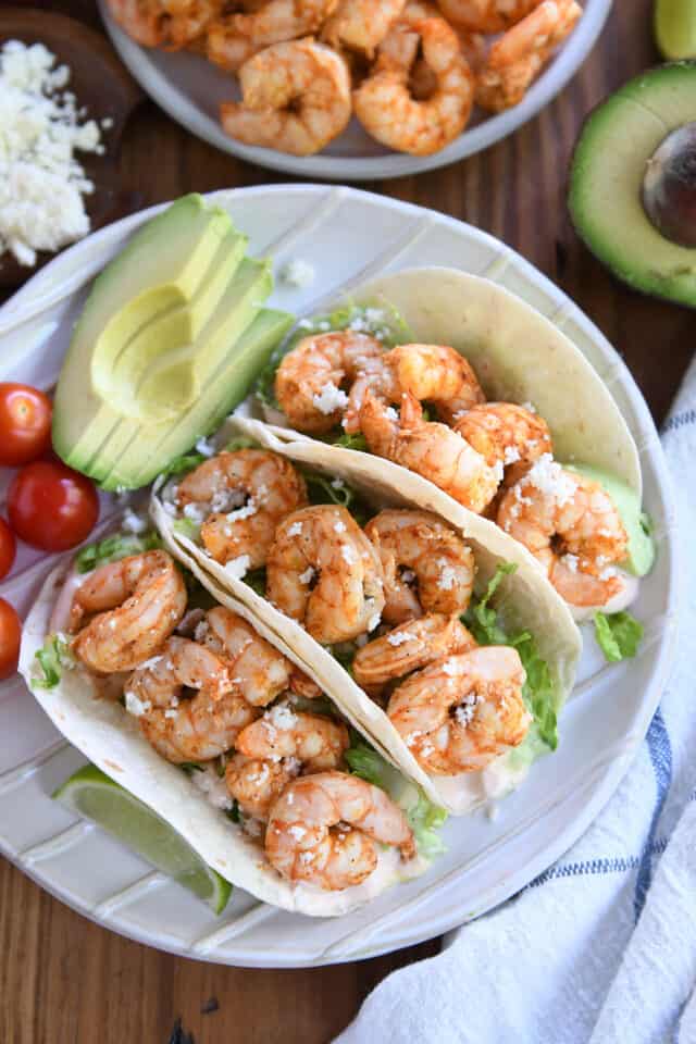 Three shrimp tacos served on a white plate with avocado and cherry tomatoes.