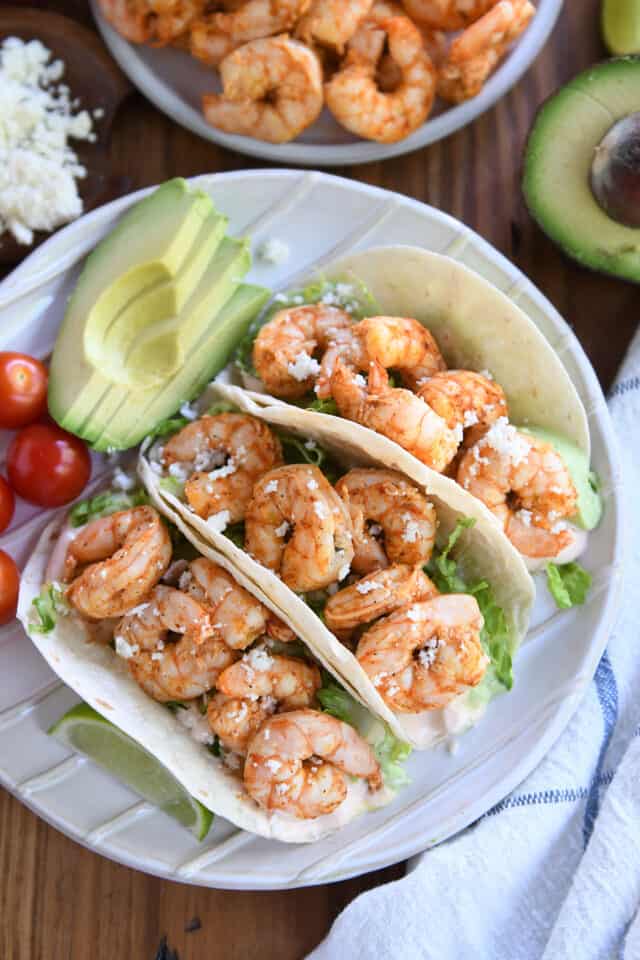 Three shrimp tacos on white plate with avocado and cherry tomato.