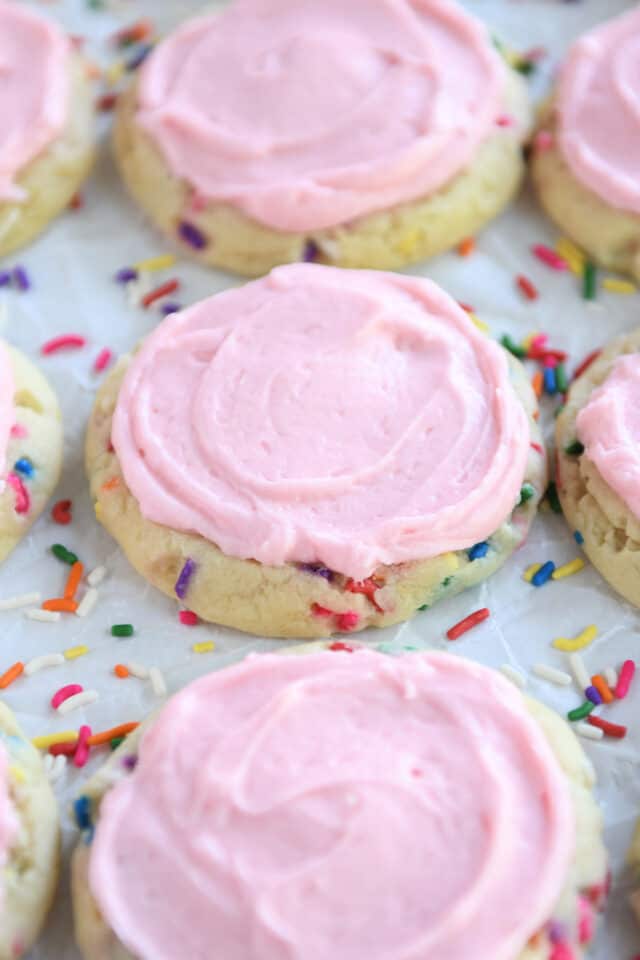 A sugar cookie with sprinkles in the dough, pink frosting on a parchment paper, and more sprinkles.