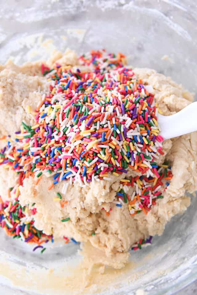 Rainbow sprinkles on top of sugar cookie dough in glass bowl.