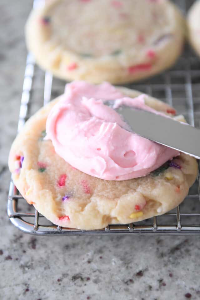 Spreading pink frosting on a sprinkle-filled sugar cookie with offset spatula.