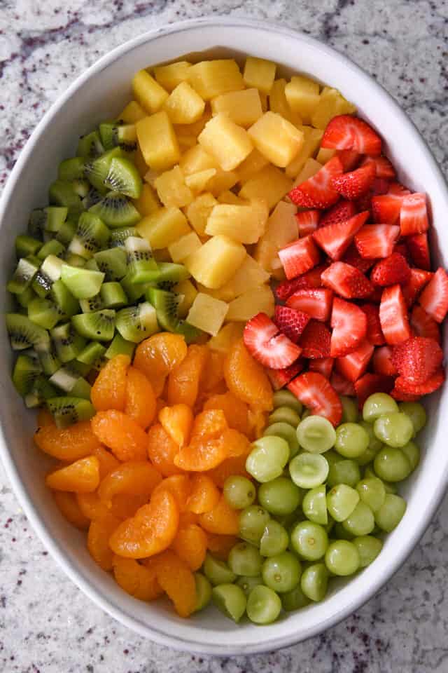 White oval bowl with grapes, mandarin oranges, kiwi fruit, pineapple and strawberries.