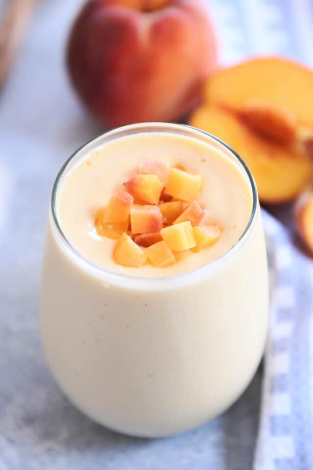 Peach orange smoothie in gl، cup with diced peaches on top and peaches in background.