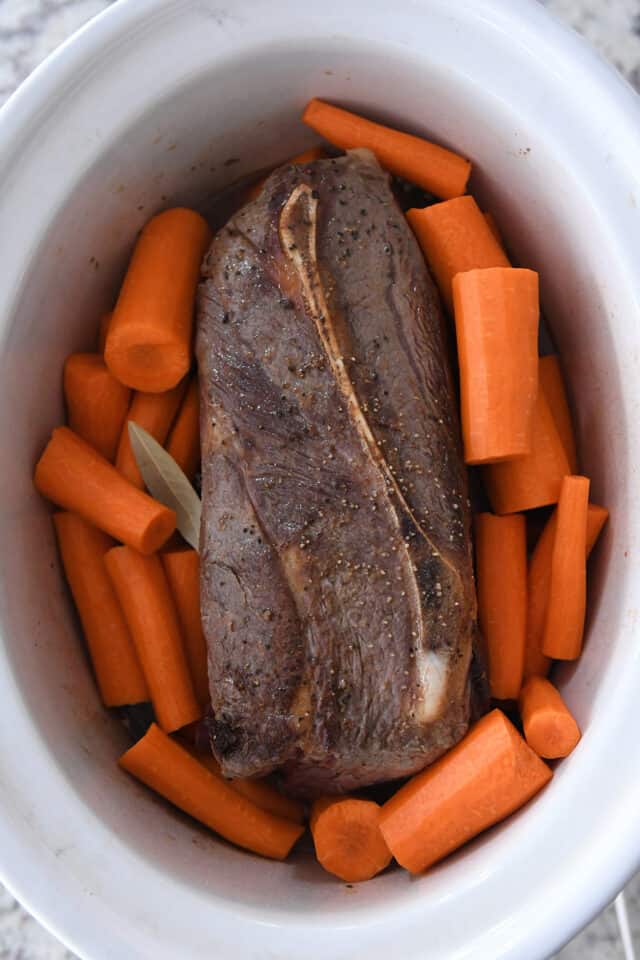 Slow cooker insert with roast, carrots and bay leaf.