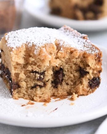 Half eaten square of chocolate chip cake dusted with powdered sugar on white plate.