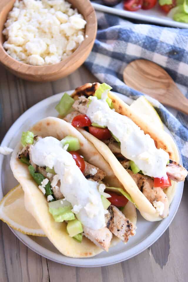 Top down view of two chicken gyros in flatbread on plate with lemon slices, tzatziki, tomatoes and lettuce.