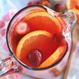 Top down view of red punch in glass pitcher with sliced oranges and strawberries.