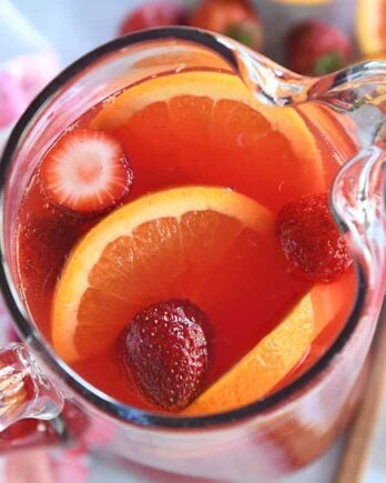 Top down view of red punch in glass pitcher with sliced oranges and strawberries.